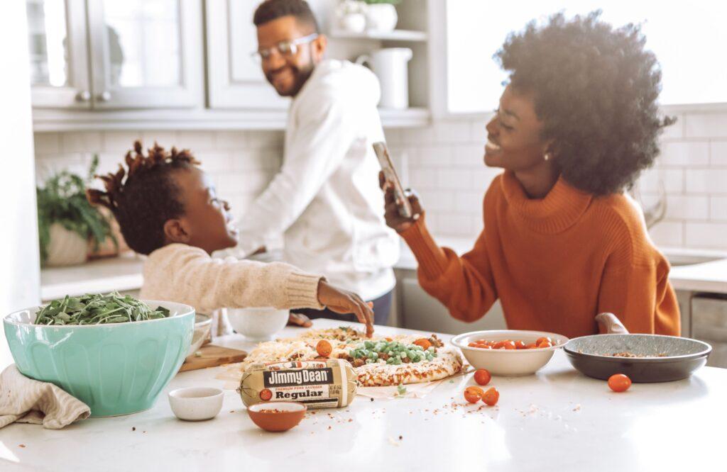 A happy family making pizza together. The mother takes a photo of her smiling toddler while Dad prepares vegetables in the background.